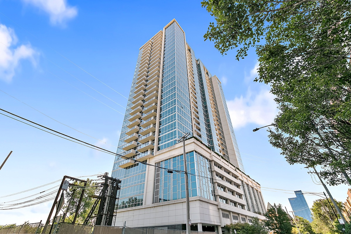 Low-angle architectural photograph of a glass residential tower framed by trees and blue sky